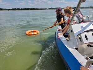 Zdjęcie przedstawia: dzieci na policyjnej łódce, które wyciągają koło ratunkowe z wody.