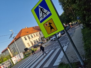 Zdjęcie przedstawia: znak drogowy przejście dla pieszych w rejonie szkoły. Z tyłu widoczny mundurowy i dzieci przechodzące przez pasy.