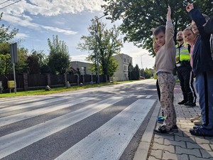 Zdjęcie przedstawia: dzieci przed przejściem dla pieszych. Dzieci stoją z ręką uniesioną do góry, policjant stoi za nimi sprawdzając czy przechodzą poprawnie przez drogę.