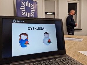 Zdjęcie przedstawia slajd na komputerze informujący o dyskusji. Z tyłu widoczny policjant.