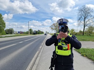 Zdjęcie przedstawia: policjanta, który stoi przy drodze i mierzy prędkość.